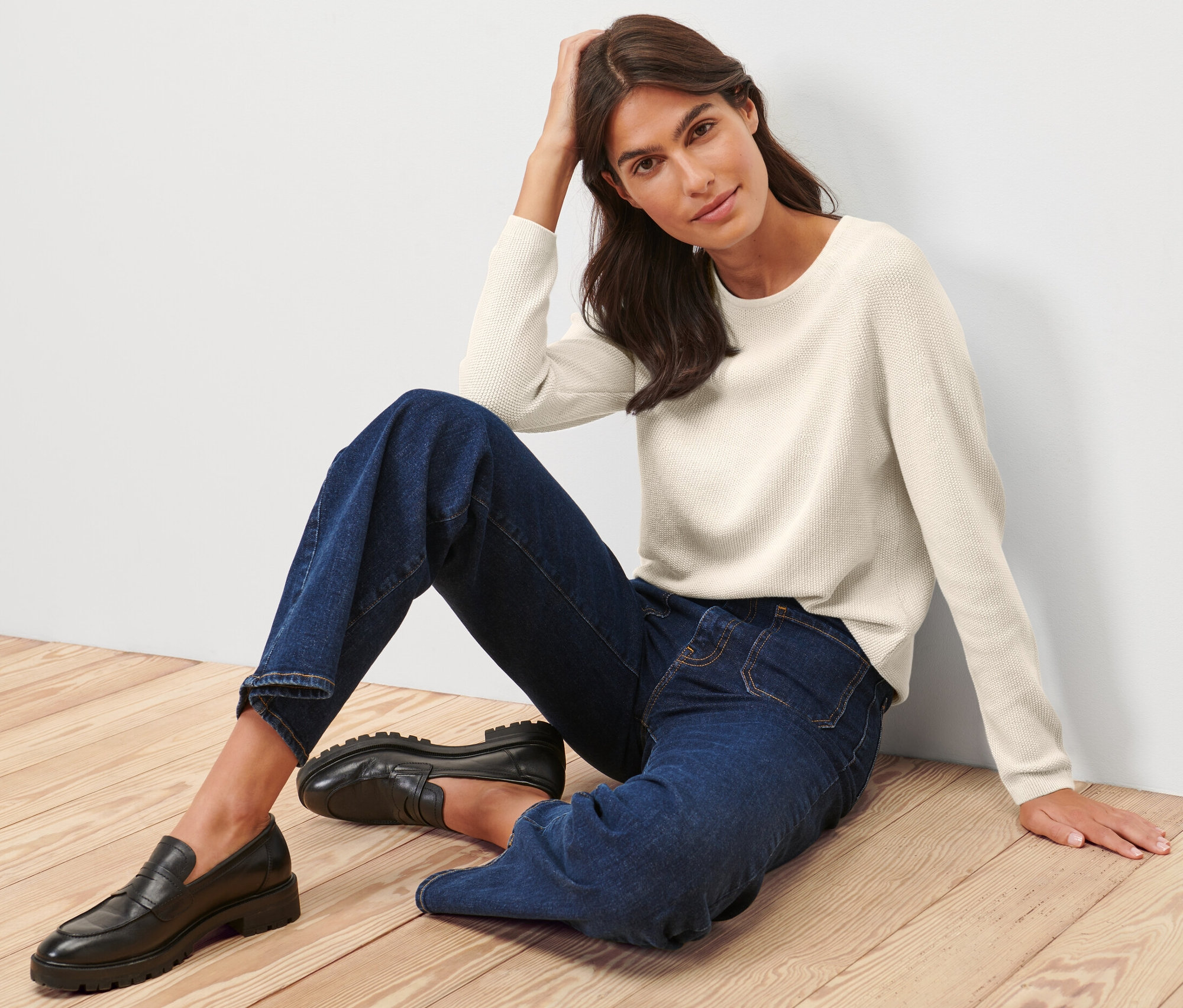 Femme assise sur un plancher en bois portant un pull en maille blanc crème.