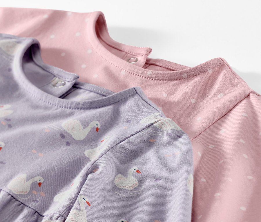 Gros plan sur deux robes en jersey pour bébé, une lilas avec motif de cygnes, l'autre rose avec des pois blancs.