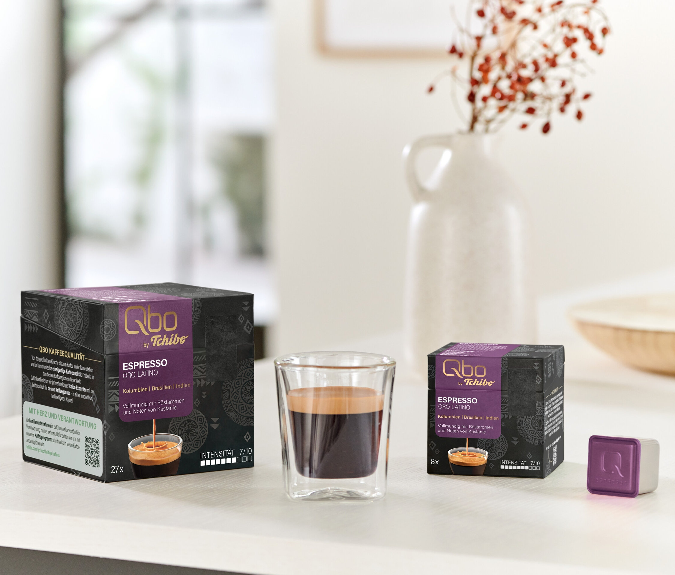 Sur une table, des paquets de capsules Qbo Espresso ORO TOLIMA (27 et 8 capsules) sont posés à côté d'un verre d'espresso et d'une capsule individuelle.