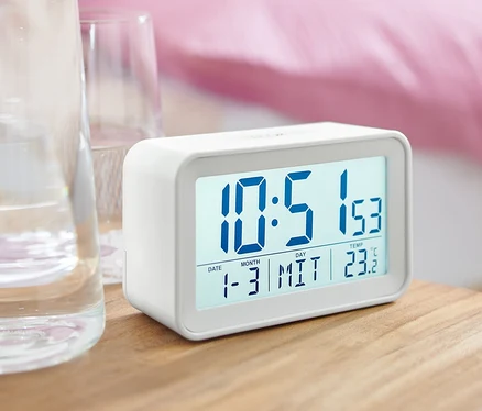 Un réveil LCD numérique blanc sur une table de chevet à côté d'un verre d'eau.