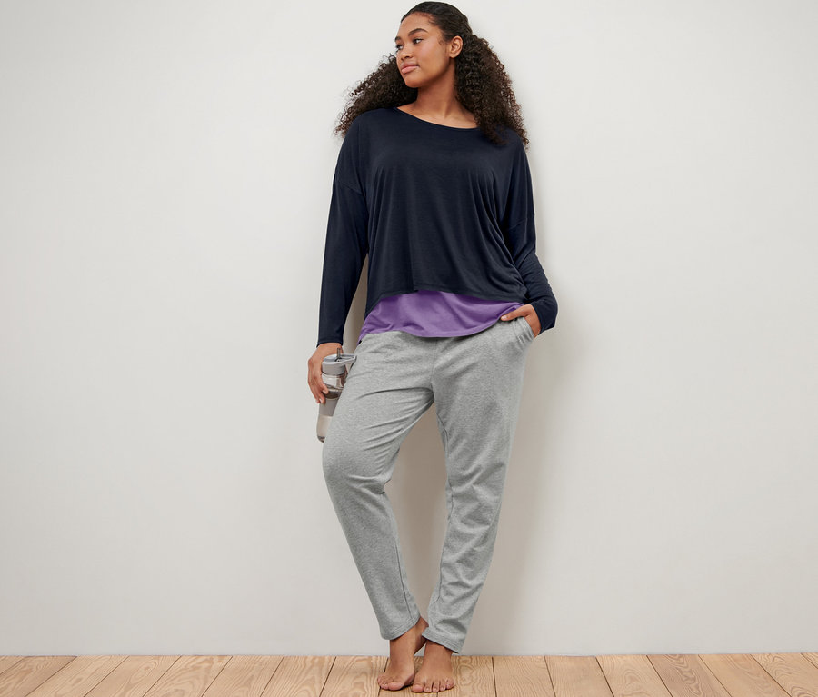 Une femme appuyée contre un mur porte un pantalon de jogging gris et un t-shirt de sport 2 en 1 bleu-lilas.