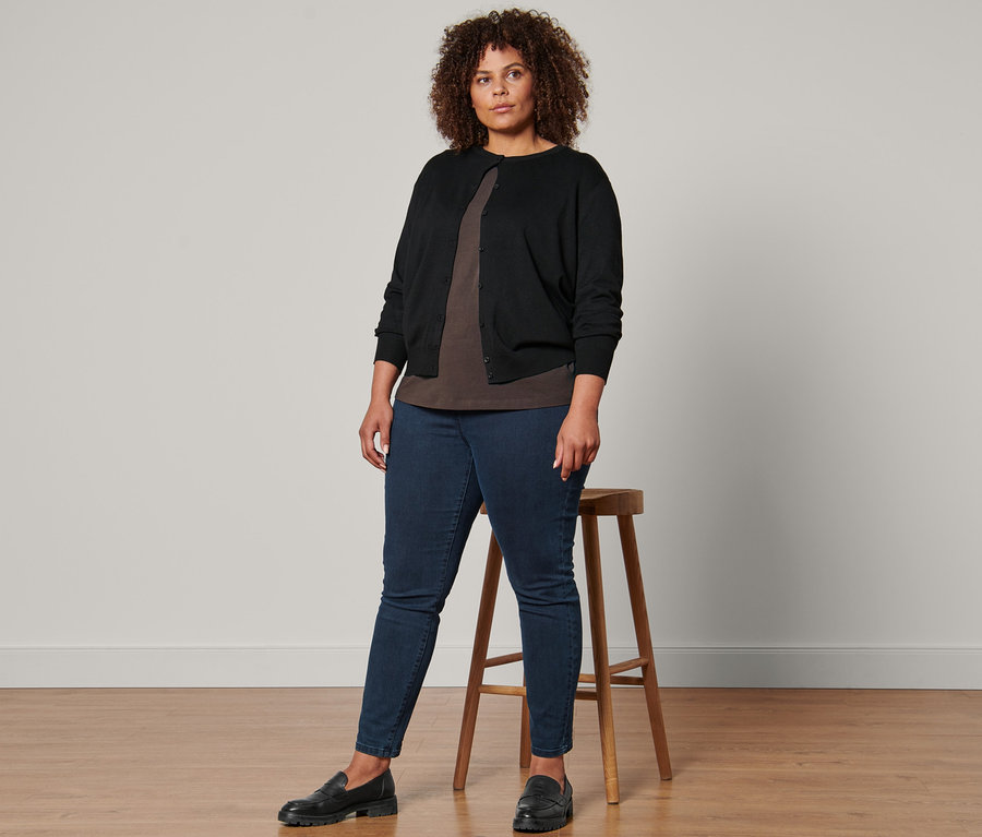 Une femme debout portant un cardigan en maille fine noir, un t-shirt manches 3/4, un tregging et des loafers en cuir.