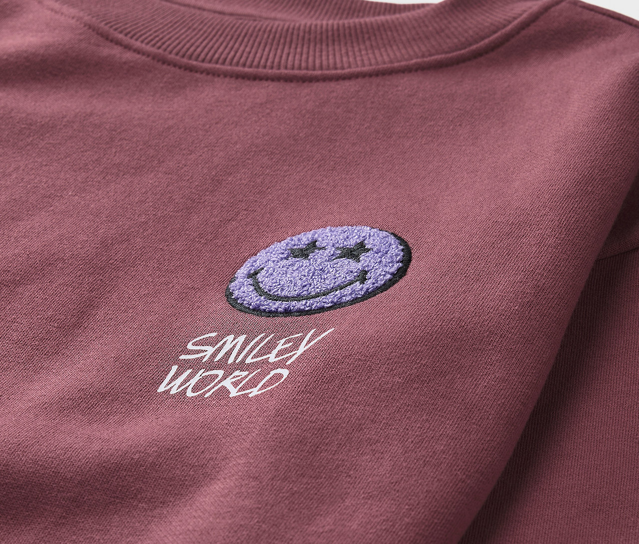 Détail d'un sweat-shirt bordeaux pour enfant SMILEYWORLD® avec un smiley violet.