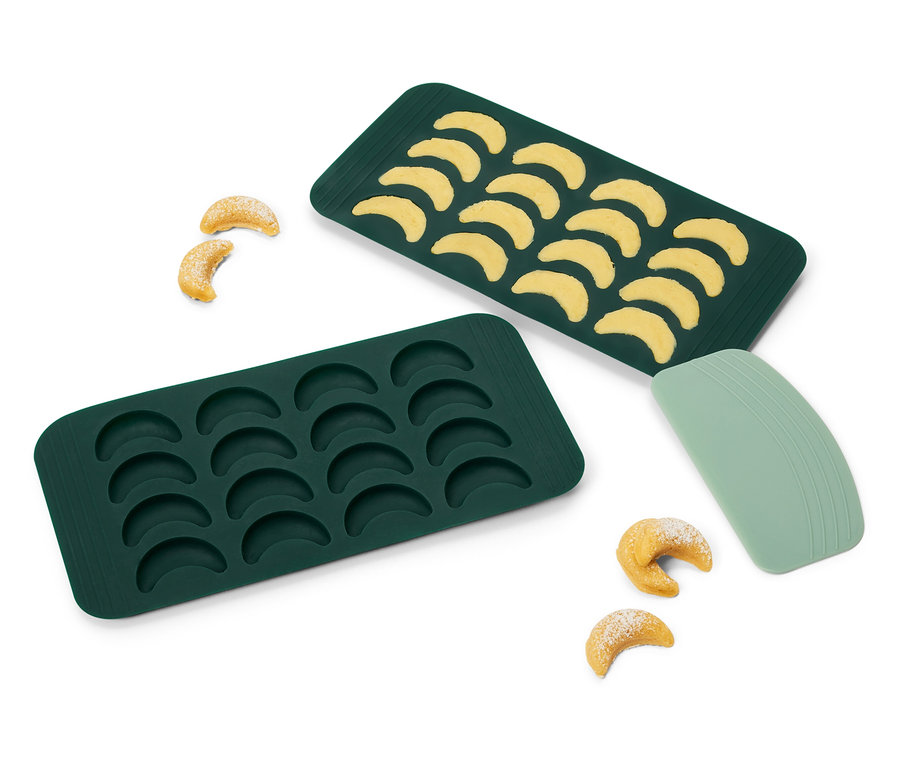 Deux moules à pâtisserie en silicone verts en forme de croissant, un rempli de pâte et deux biscuits cuits à côté.
