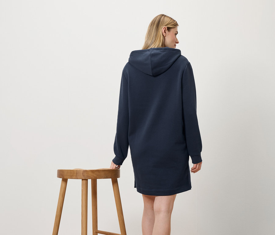 Une femme en robe à capuche bleu marine se tient à côté d'un tabouret en bois.
