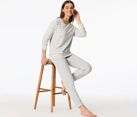 Femme souriante portant un ensemble pyjama en tissu éponge SCHIESSER rayé gris et blanc, assise sur un tabouret en bois.