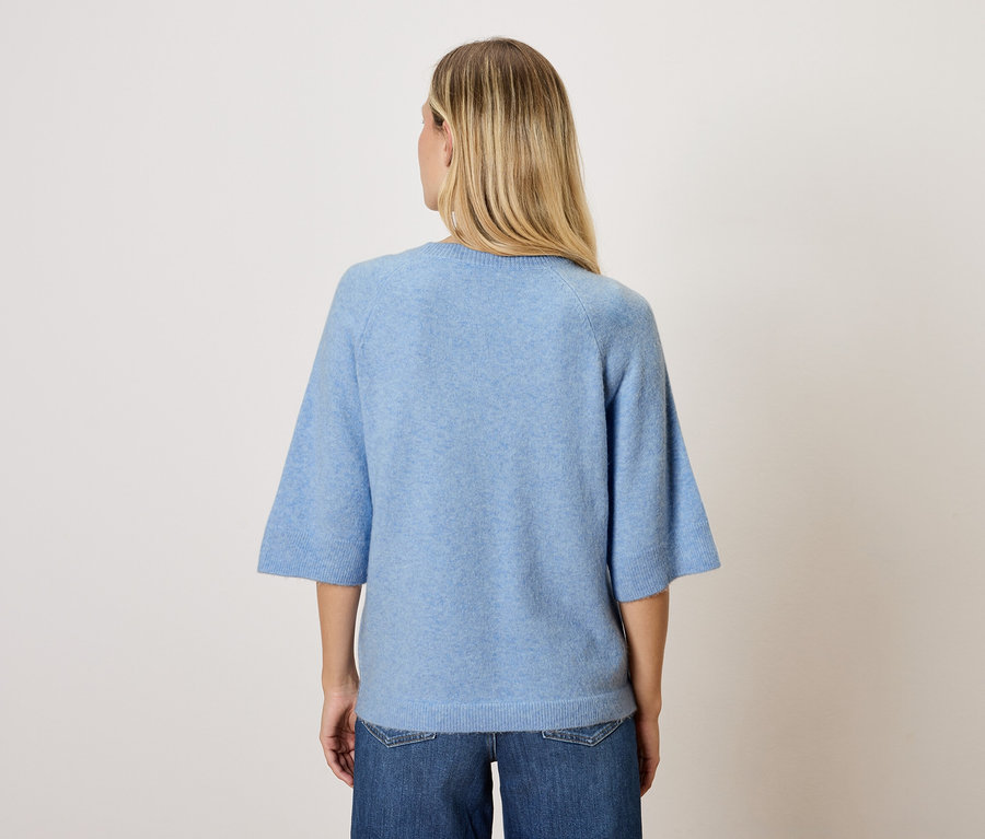 Une femme blonde se tient dos à la caméra, portant un haut bleu et un jean.