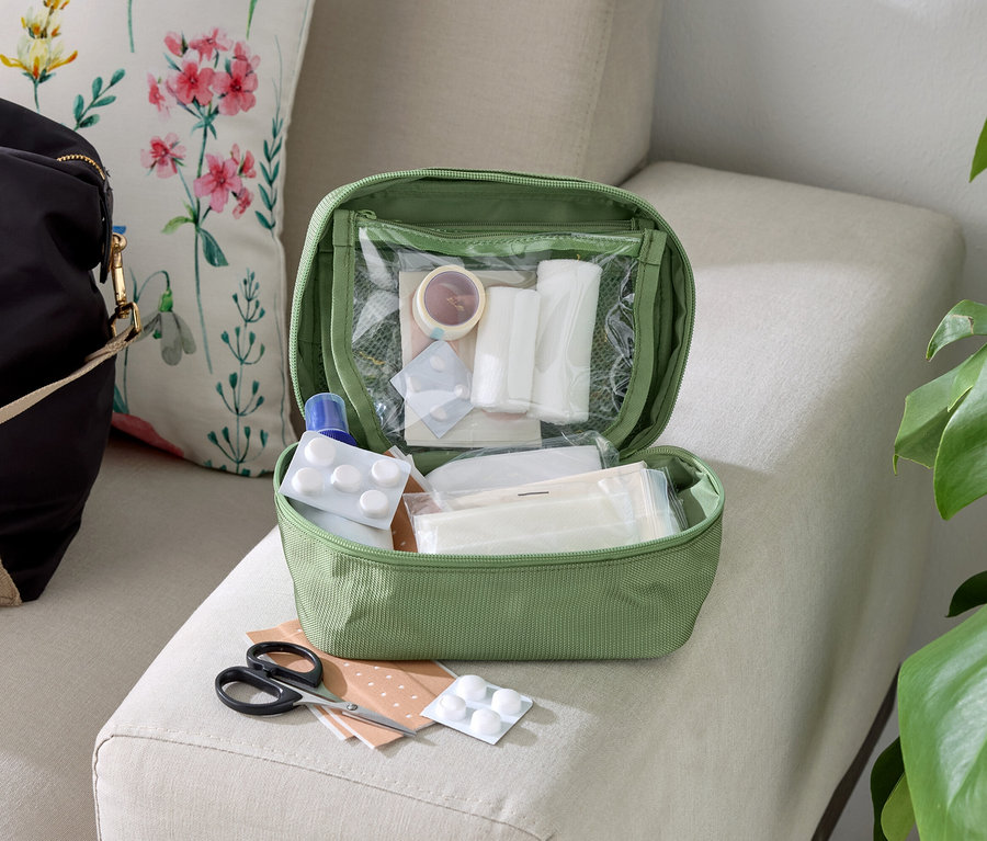 Une trousse de premiers secours verte ouverte avec son contenu sur un canapé beige à côté d'un sac et d'un coussin.