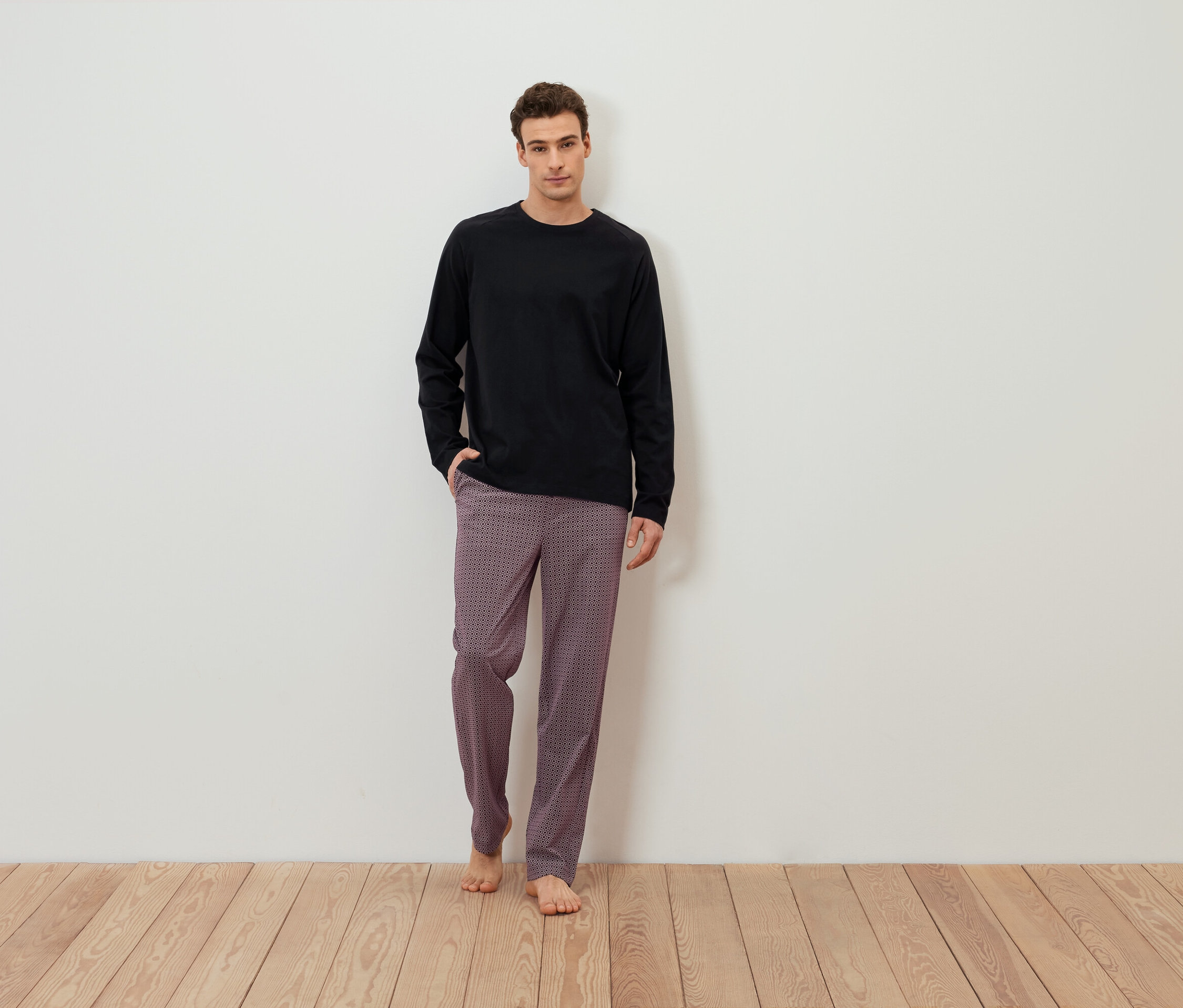 Un homme se tient pieds nus devant un mur blanc, portant un t-shirt noir à manches longues et un pantalon à motifs.