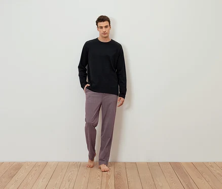 Un homme se tient pieds nus devant un mur blanc, portant un t-shirt noir à manches longues et un pantalon à motifs.