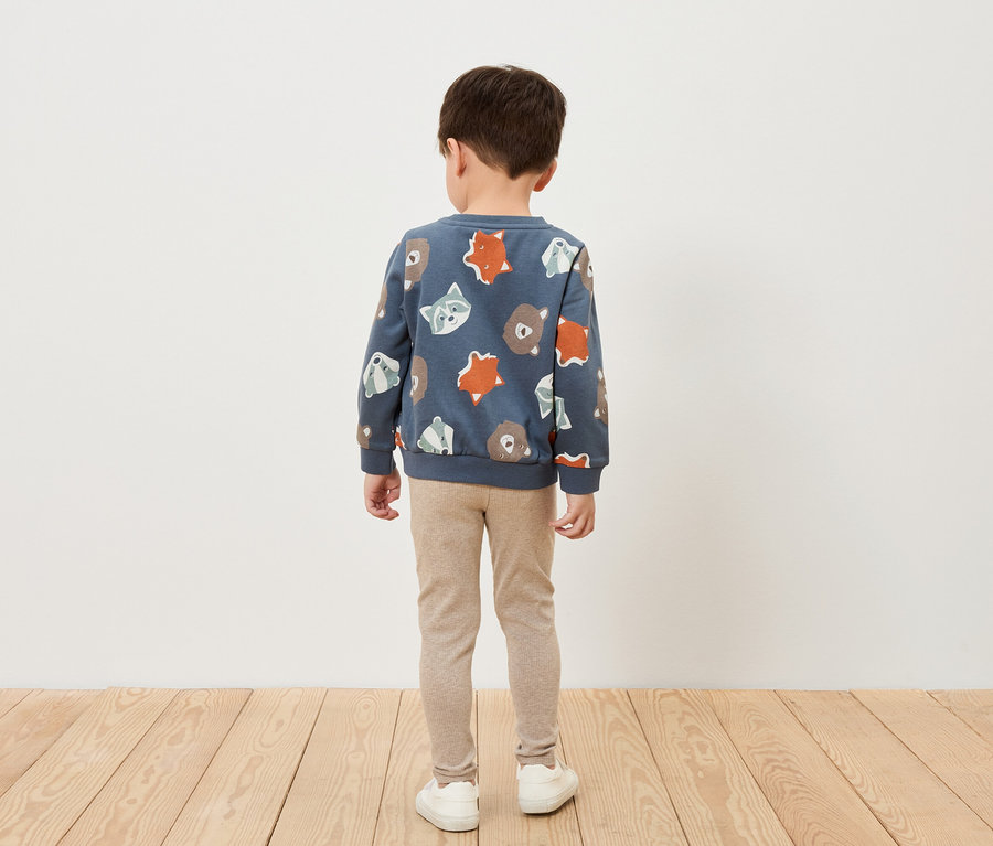 Un garçon se tient dos à la caméra, portant un sweat-shirt bleu à motifs d'animaux, un legging beige et des chaussures blanches.