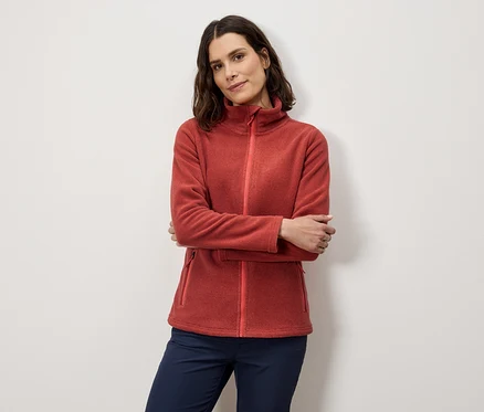 Femme posant les bras croisés portant une veste polaire orange et un pantalon bleu marine.