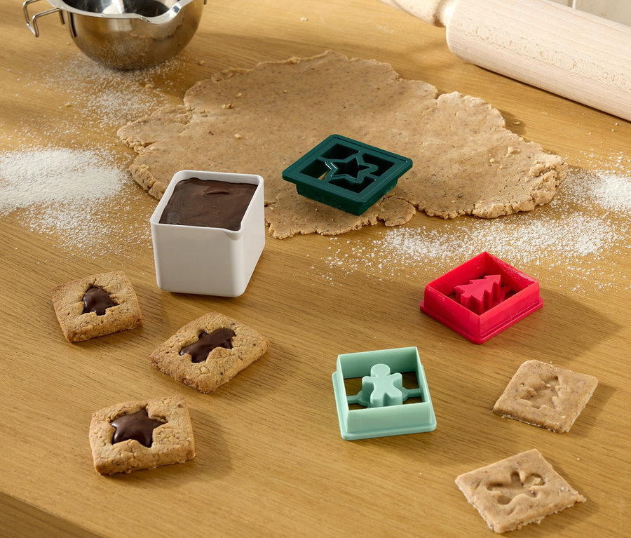 Sur une table en bois se trouvent de la pâte étalée, des emporte-pièces, des biscuits finis et un bol de glaçage.