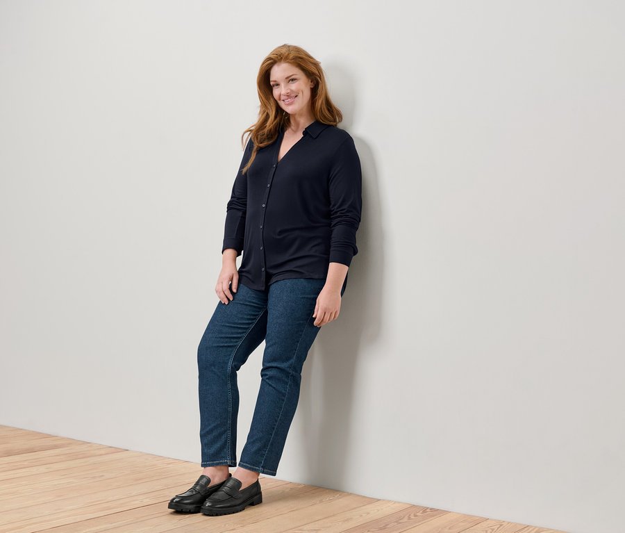 Une femme souriante aux longs cheveux roux, vêtue d'une chemise bleu foncé, d'un jean et de mocassins noirs, s'appuie contre un mur blanc sur un plancher en bois.