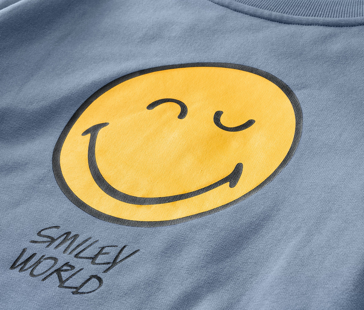 Gros plan sur un sweatshirt bleu pour enfant SMILEYWORLD® avec un smiley jaune.