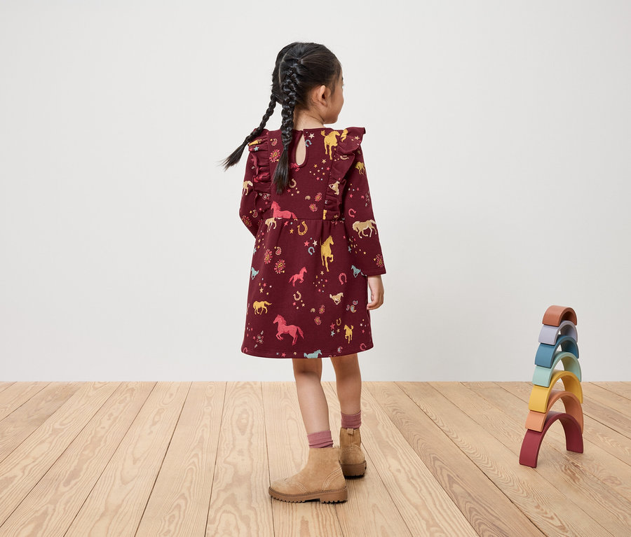 Une fille se tient dos tourné, portant une robe bordeaux avec un imprimé de chevaux, et des jouets.