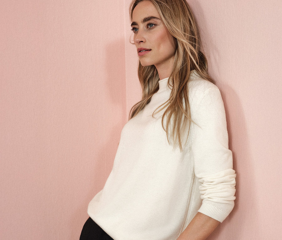 Une femme appuiée contre un mur rose porte un pull en tricot beige à bord roulé.