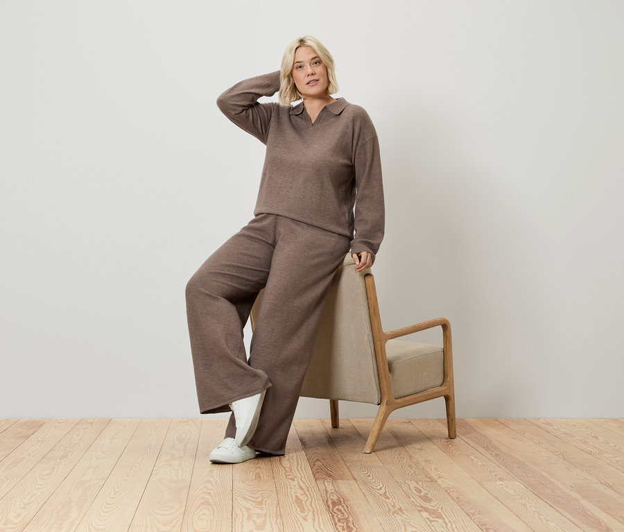 Une femme blonde portant un pull marron et un pantalon large pose, appuyée sur une chaise.