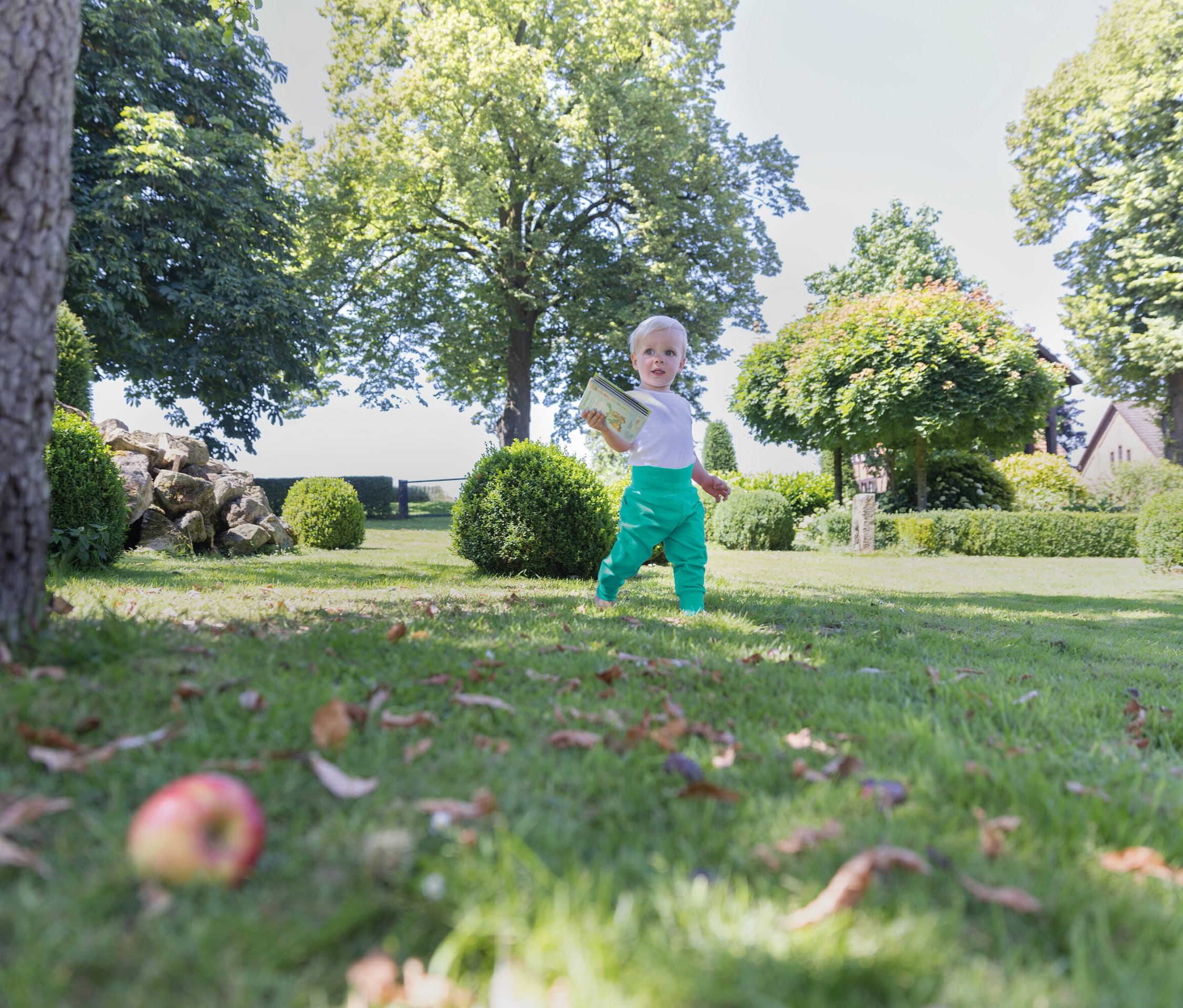 Un bébé vêtu d'un pantalon vert marche sur l'herbe dans un jardin.