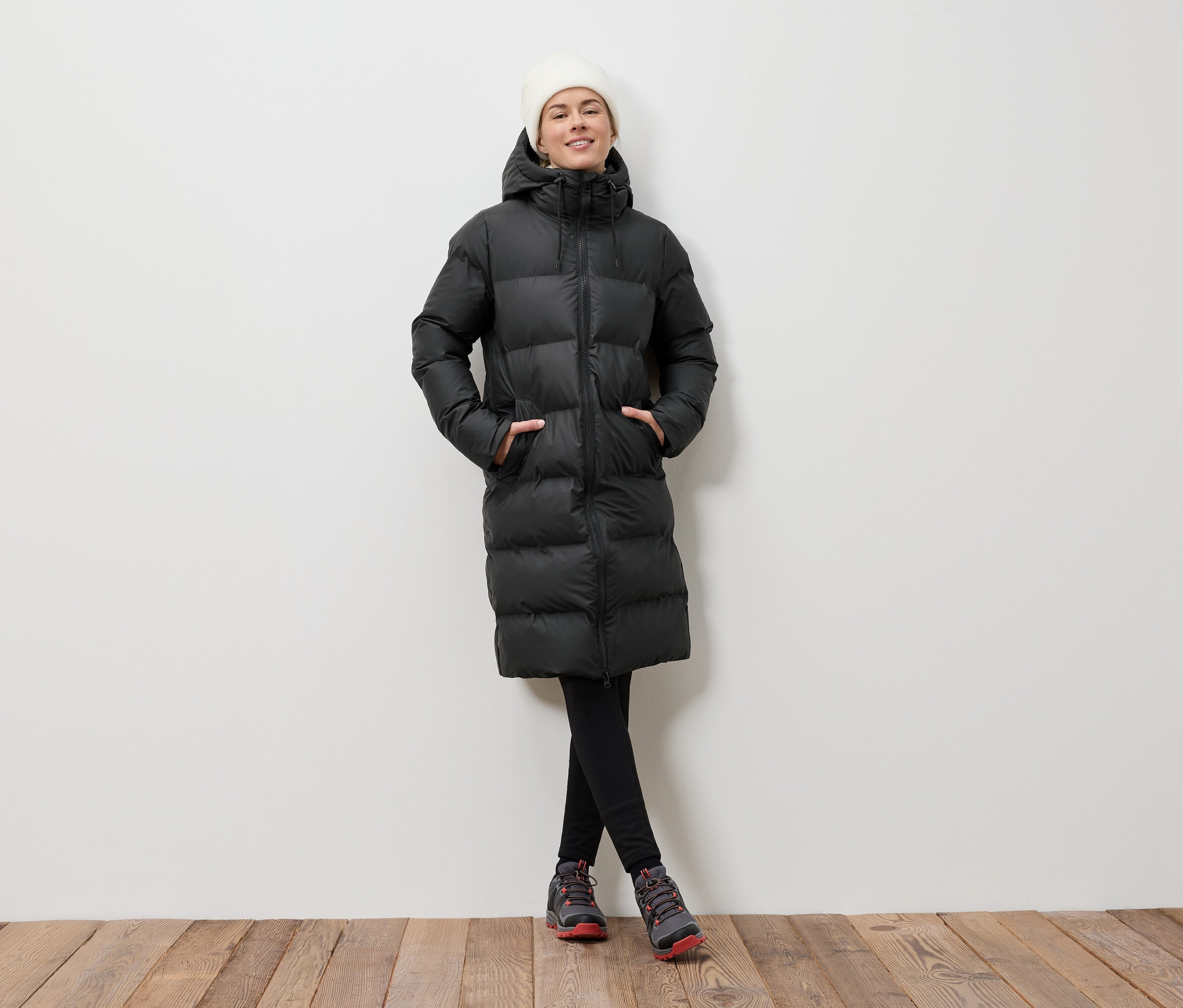 Une femme pose sur un plancher en bois, portant une doudoune noire et un bonnet blanc.