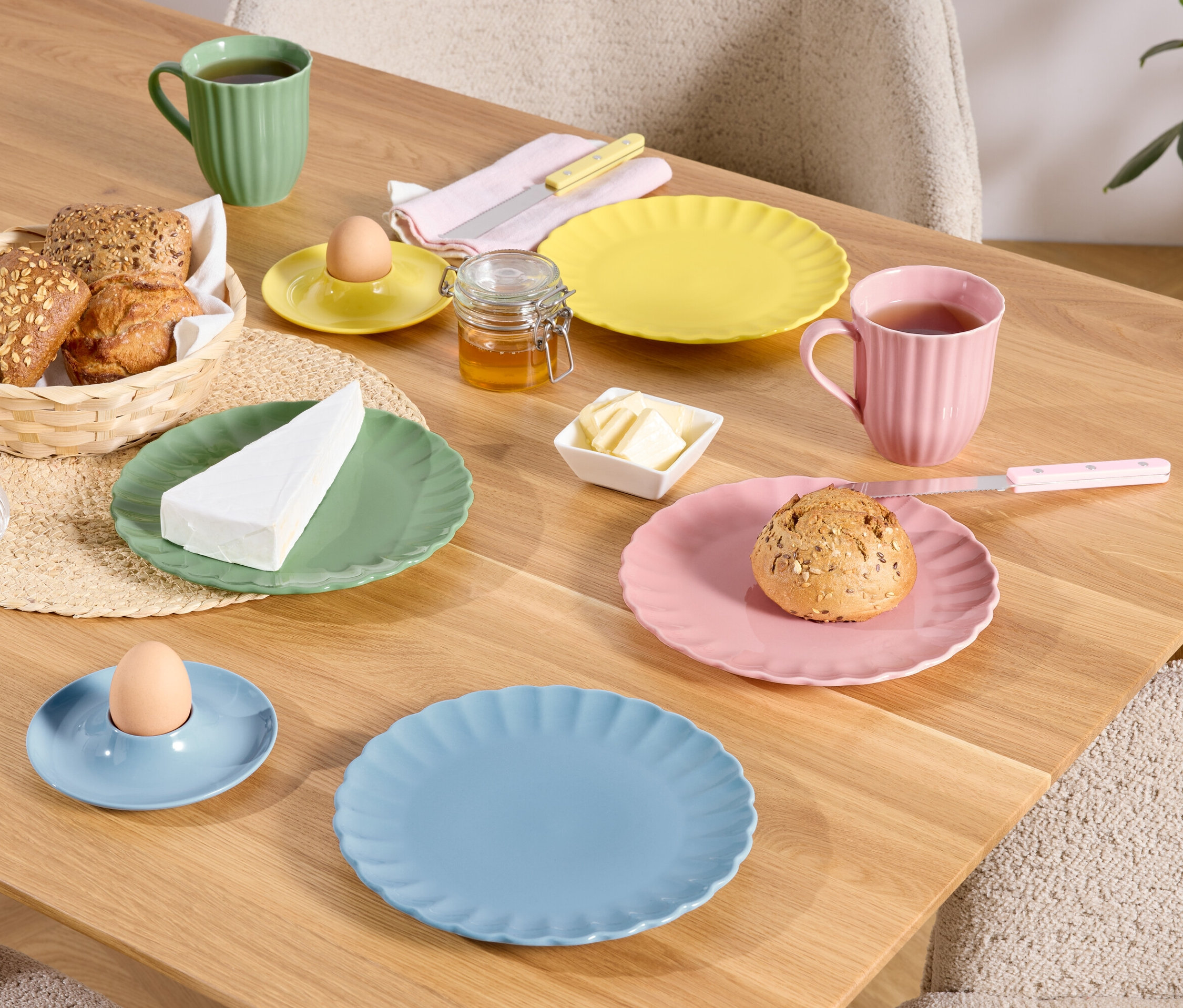 Une table en bois est dressée avec des assiettes colorées, des tasses et des bols contenant de la nourriture, notamment du pain dans un panier, un œuf dans un coquetier et un morceau de fromage.