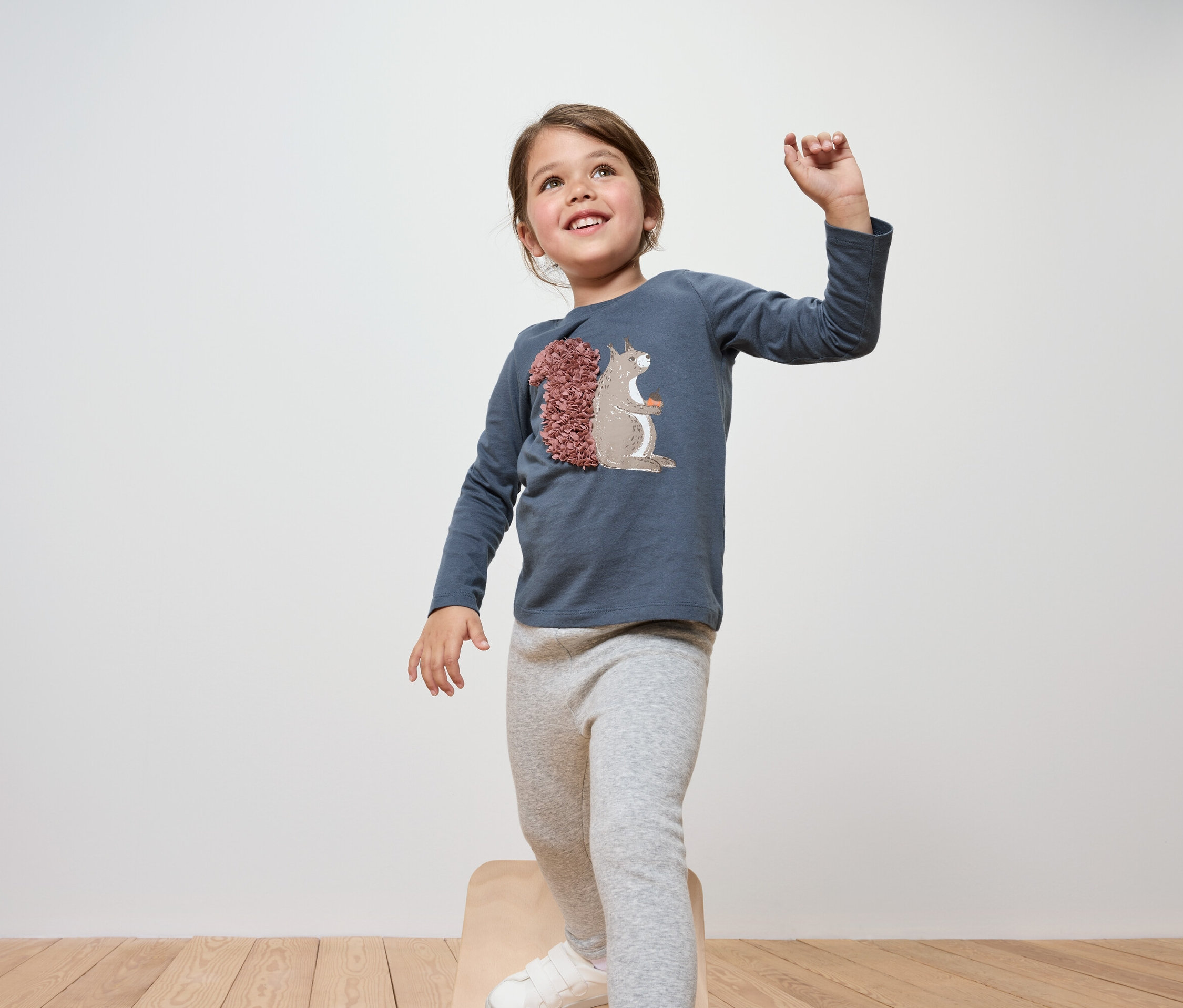 Une fillette souriante se tient sur un tabouret en bois, portant un t-shirt bleu à manches longues et un legging gris.