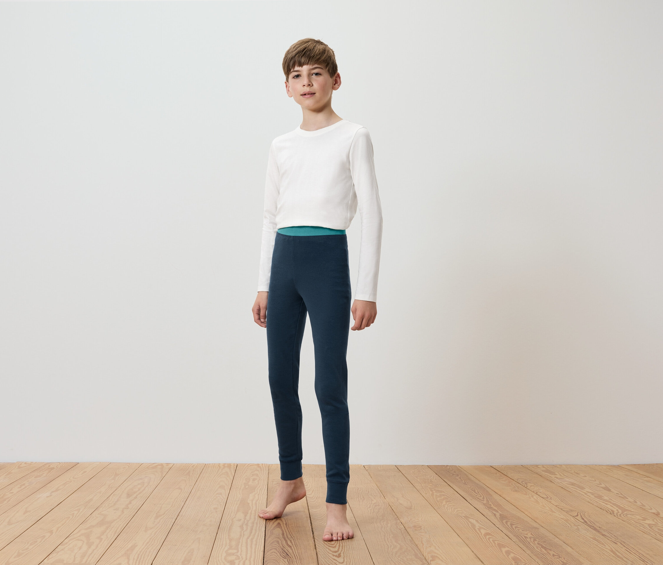 Un garçon portant un t-shirt blanc à manches longues et un legging bleu marine se tient pieds nus sur un plancher en bois devant un mur blanc.