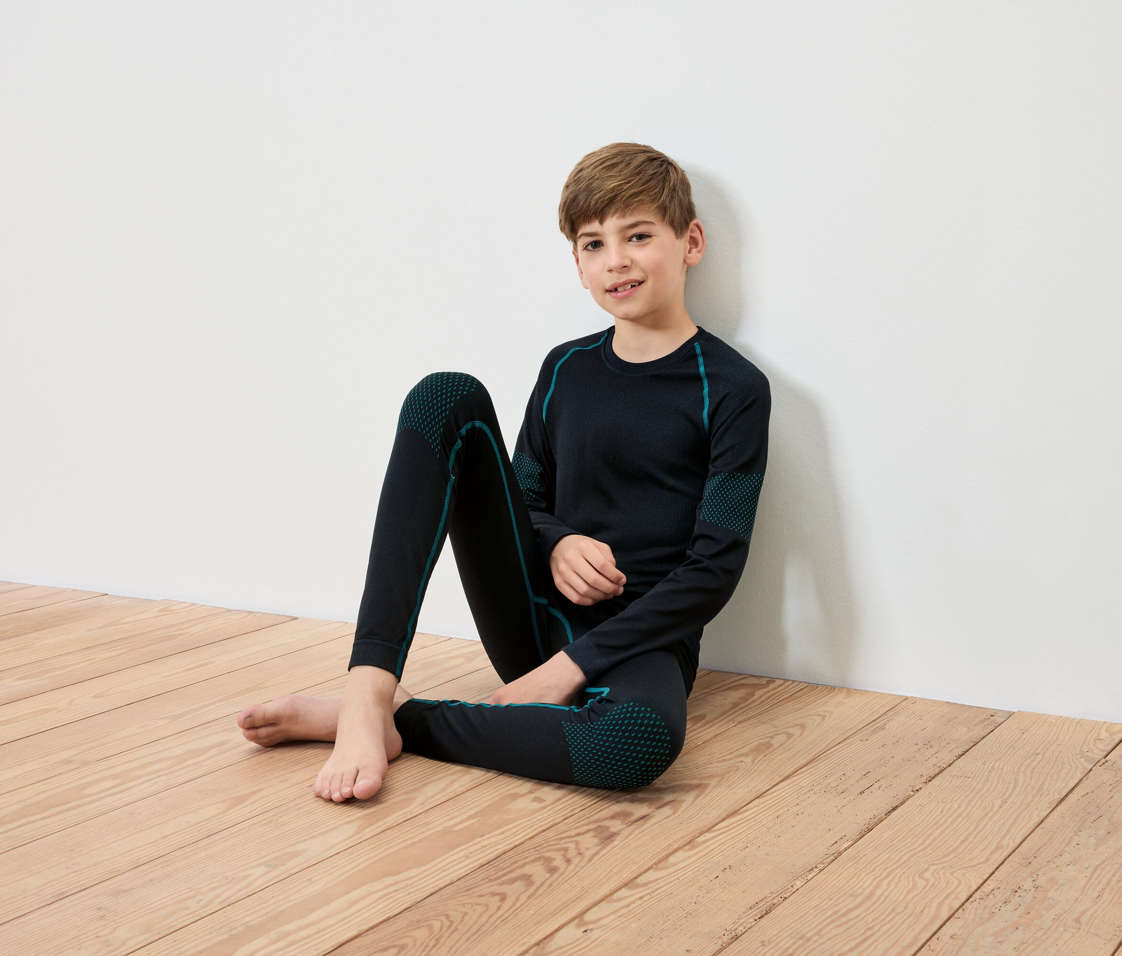 Un garçon est assis sur un plancher en bois, adossé à un mur blanc. Il porte des sous-vêtements thermiques noirs avec des détails turquoise.