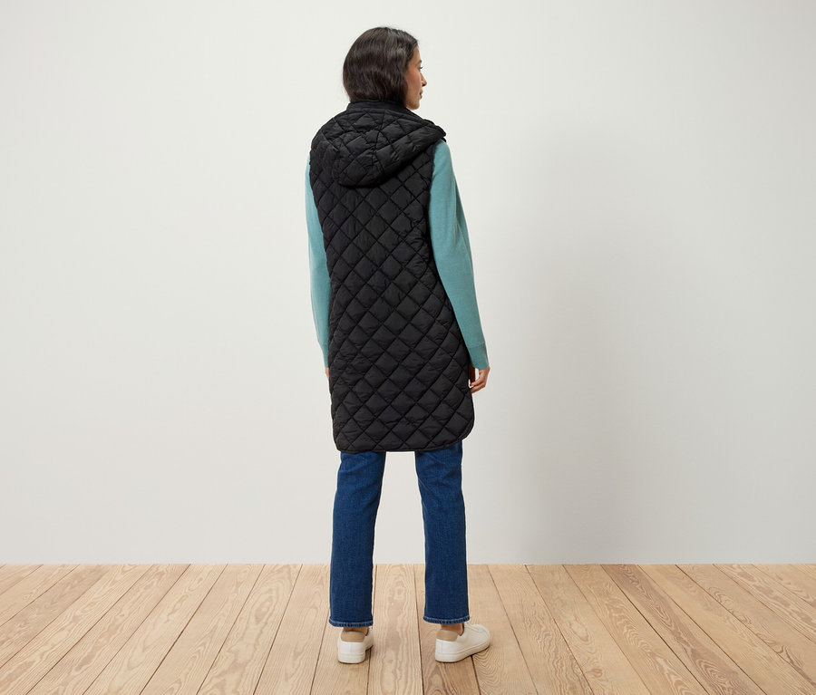 Une femme, vue de dos, porte un long gilet noir matelassé à capuche, un pull bleu et un jean.