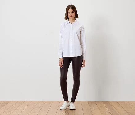 Femme en chemise blanche et leggings marron debout sur un plancher en bois.