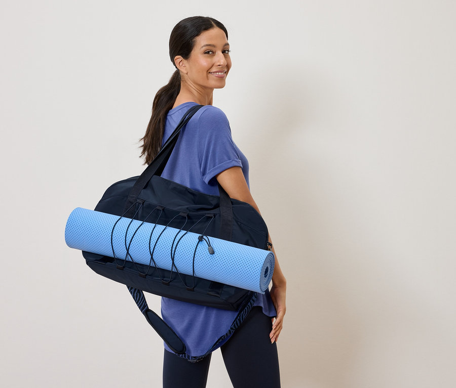 Femme portant un tapis de yoga bleu dans un sac de sport noir sur l'épaule et regardant par-dessus son épaule.