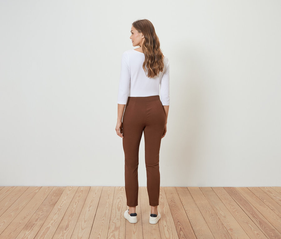 Une femme se tient dos à la caméra et porte un pantalon marron et un chemisier blanc.