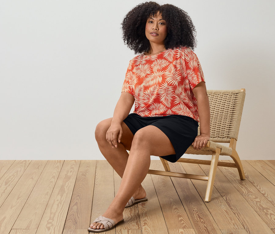 Une femme est assise sur une chaise et porte une blouse imprimée et un short en jersey noir.