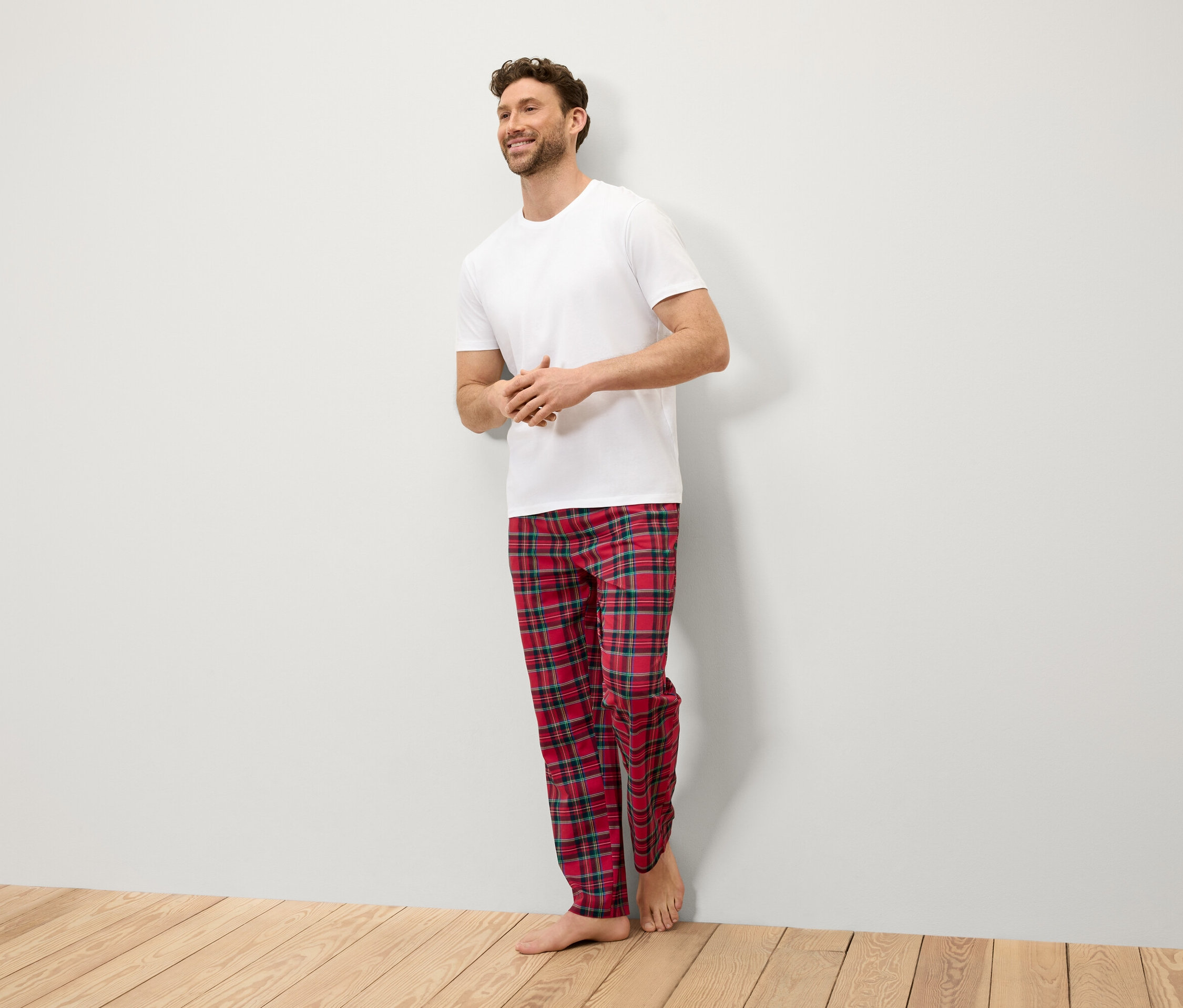 Un homme souriant se tient contre un mur, vêtu d'un t-shirt blanc et d'un pantalon à carreaux rouge.