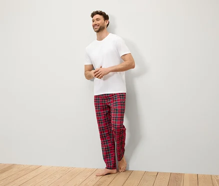 Un homme souriant se tient contre un mur, vêtu d'un t-shirt blanc et d'un pantalon à carreaux rouge.