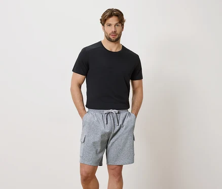 Un homme les mains dans les poches d'un short gris à cordon de serrage et d'un t-shirt noir à manches courtes.