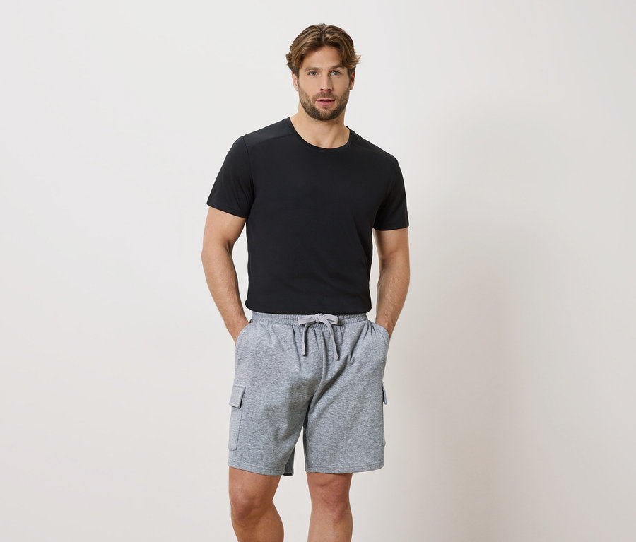 Un homme les mains dans les poches d'un short gris à cordon de serrage et d'un t-shirt noir à manches courtes.