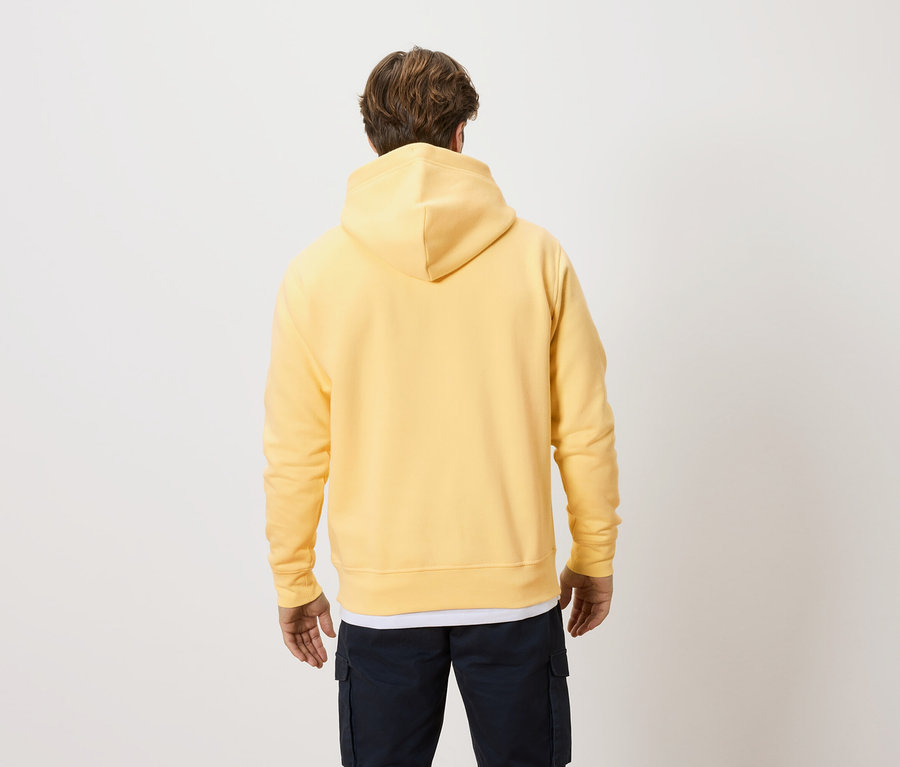 Vue arrière d'un homme blond portant un sweat à capuche jaune et un pantalon foncé sur un fond clair.