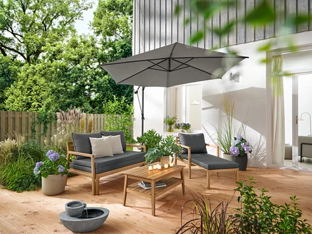 Sur une terrasse en bois se trouvent un fauteuil de jardin extensible, un canapé de jardin extensible, une table basse de jardin d'environ 80 x 50 cm et une fontaine de jardin solaire. Un parasol Leco anthracite crée de l'ombre. 2 coussins d’extérieur avec texture lin et 6 bougies LED rechargeables d’extérieur décorent l'espace.