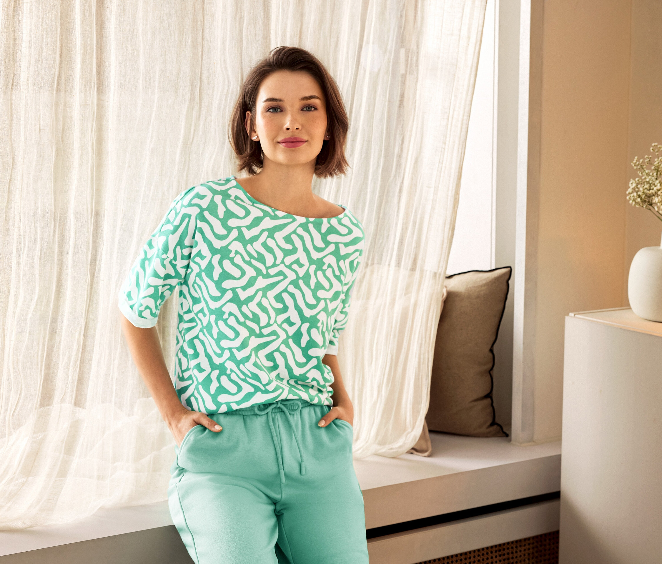 Femme portant un pantalon vert et un haut à motifs posant près d'une fenêtre. On aperçoit des puces d’oreille avec arc en zircons en argent 925 et un vase en arrière-plan.