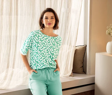 Femme portant un pantalon vert et un haut à motifs posant près d'une fenêtre. On aperçoit des puces d’oreille avec arc en zircons en argent 925 et un vase en arrière-plan.