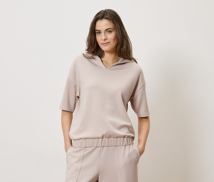 Une femme aux cheveux bruns porte un haut à capuche beige et un pantalon assorti sur un fond uni.