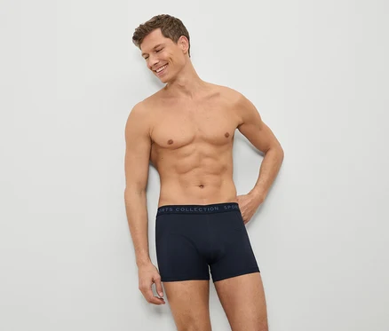 Un homme torse nu en boxer bleu appuyé contre un mur blanc.