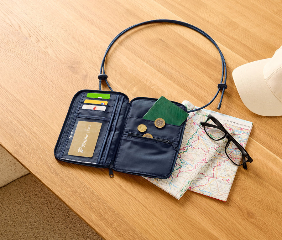 Portefeuille de voyage bleu marine ouvert avec des cartes, un passeport et des pièces de monnaie sur une table en bois, avec une carte, des lunettes noires et une casquette blanche.