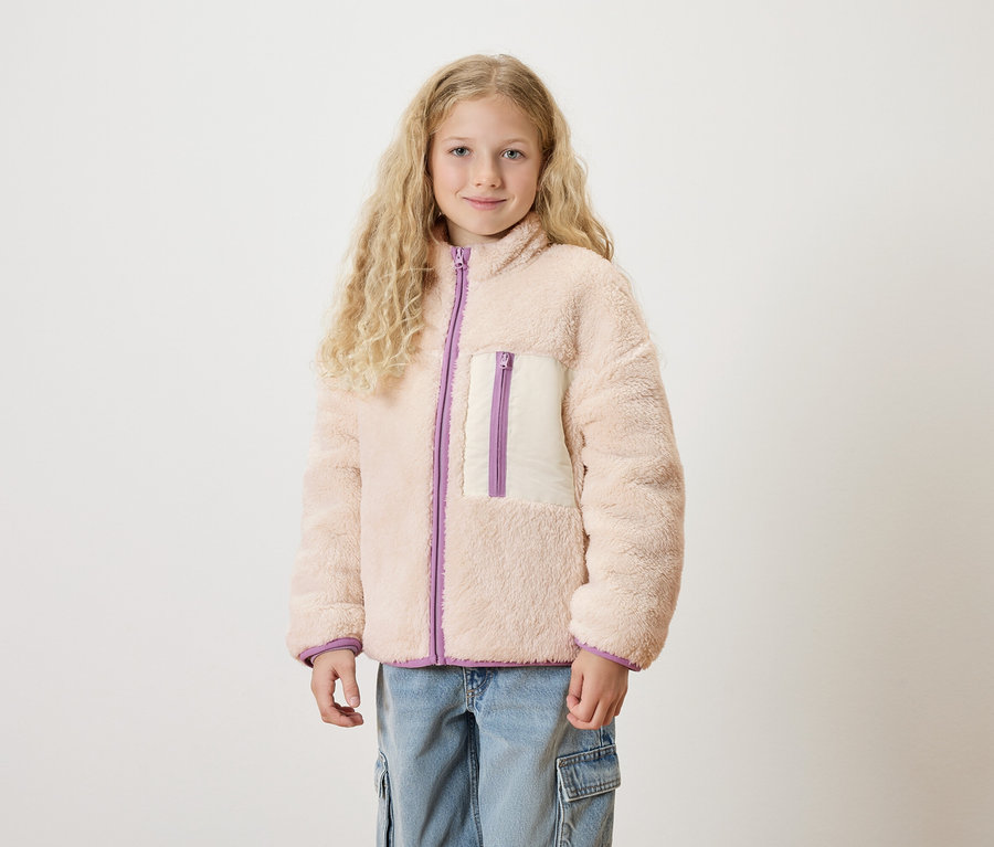 Une jeune fille aux cheveux blonds et bouclés porte une veste teddy crème avec des détails violets et un jean.