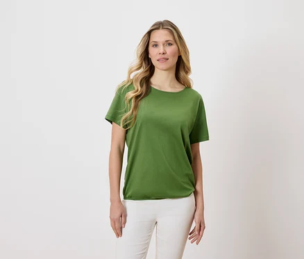 Une femme aux cheveux blonds et ondulés porte un t-shirt vert et un pantalon blanc sur un fond clair.