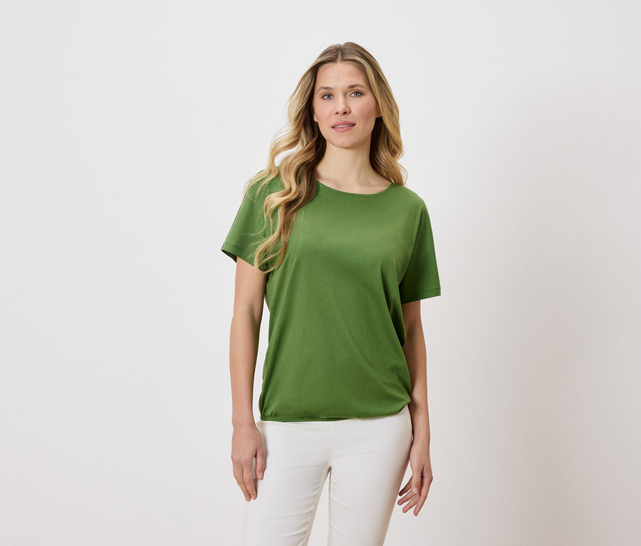 Une femme aux cheveux blonds et ondulés porte un t-shirt vert et un pantalon blanc sur un fond clair.