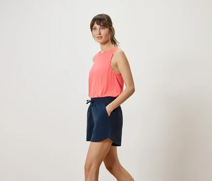 Femme portant un débardeur corail et un short bleu marine, la main dans la poche, vue de côté.