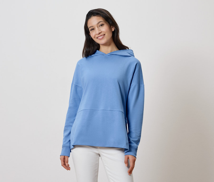 Femme aux longs cheveux foncés portant un sweat à capuche bleu clair et un pantalon blanc.