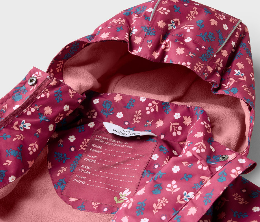 Gros plan d'une veste pour enfant bordeaux à capuche avec un motif floral, doublée de polaire rose à l'intérieur. Une étiquette avec un espace pour le nom et le numéro de téléphone est visible.